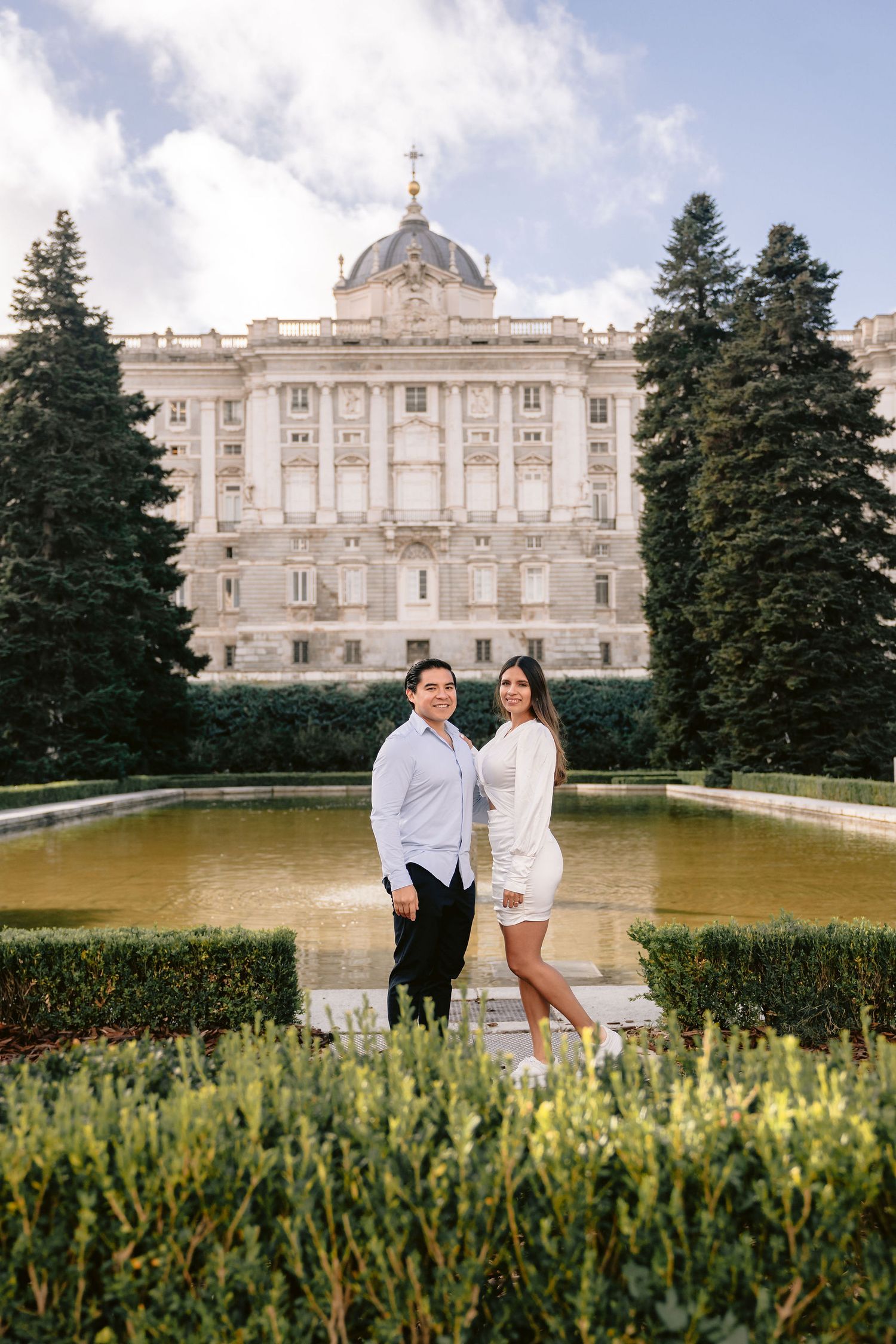 A couple poses in front of the Royal Palace of Madrid with its majestic facade and reflecting pool.