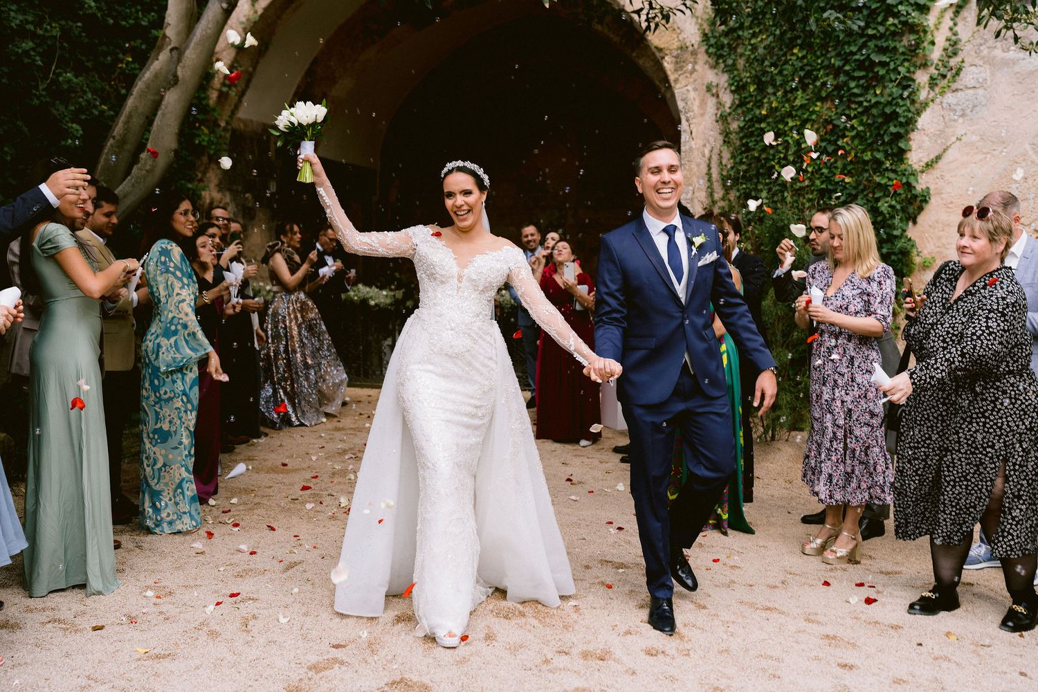 Newlyweds celebrate while exiting their wedding ceremony as guests toss confetti and cheer along a tree-lined path.