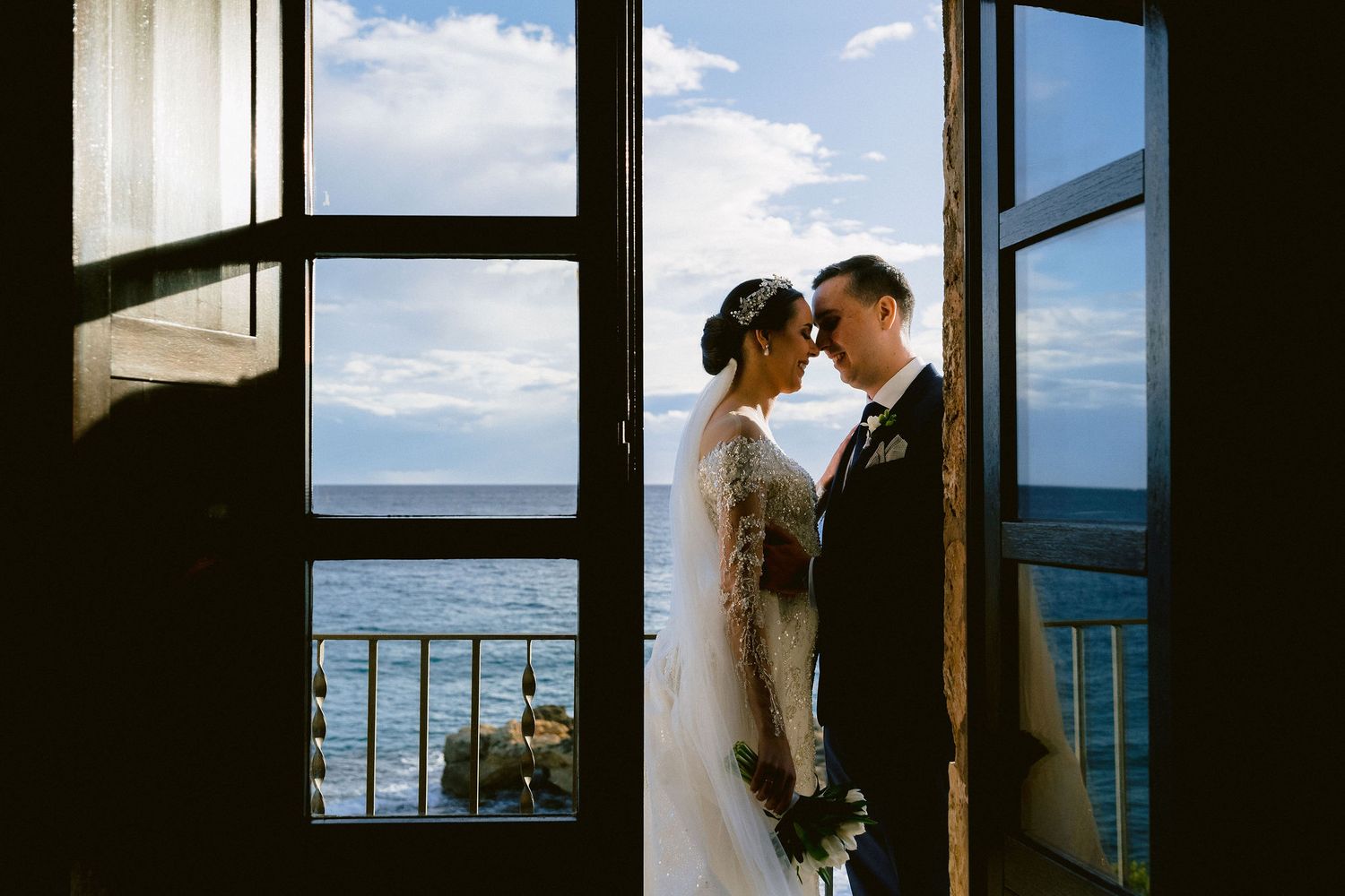 A romantic couple embraces by a window overlooking a scenic ocean view in silhouette.