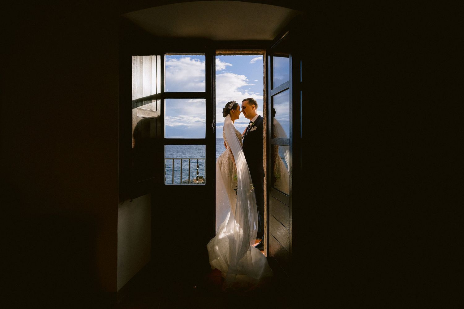 Silhouetted couple share an intimate moment by a bright window overlooking the ocean on their wedding day.