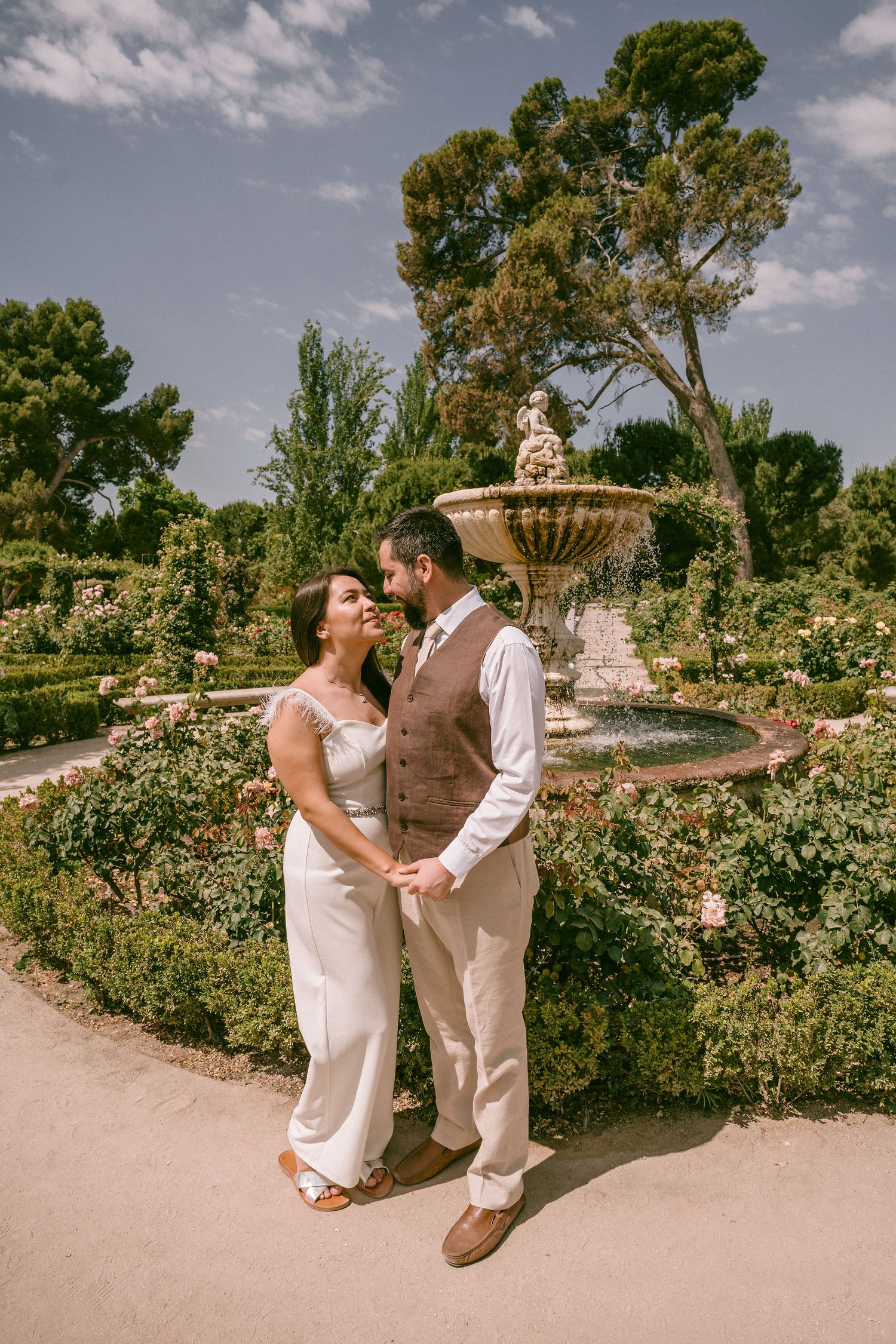 A couple in elegant attire shares a romantic moment in a garden with fountain.