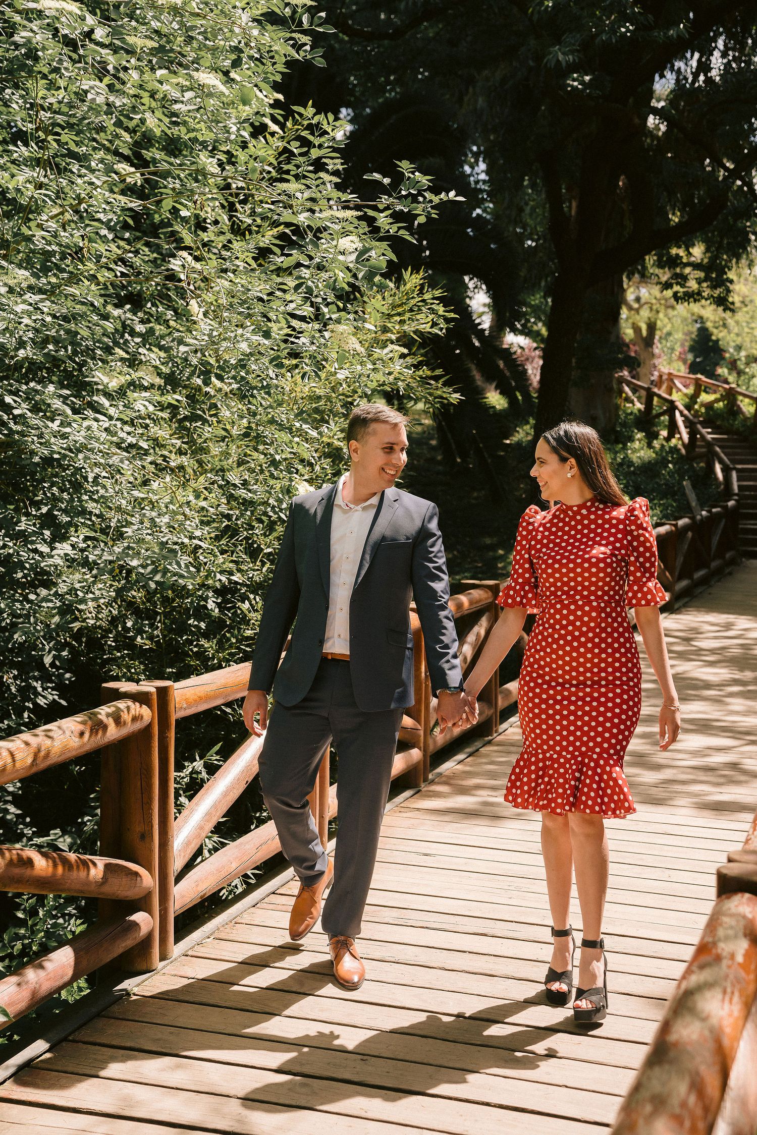 A couple walks hand in hand across a wooden bridge surrounded by lush green trees.