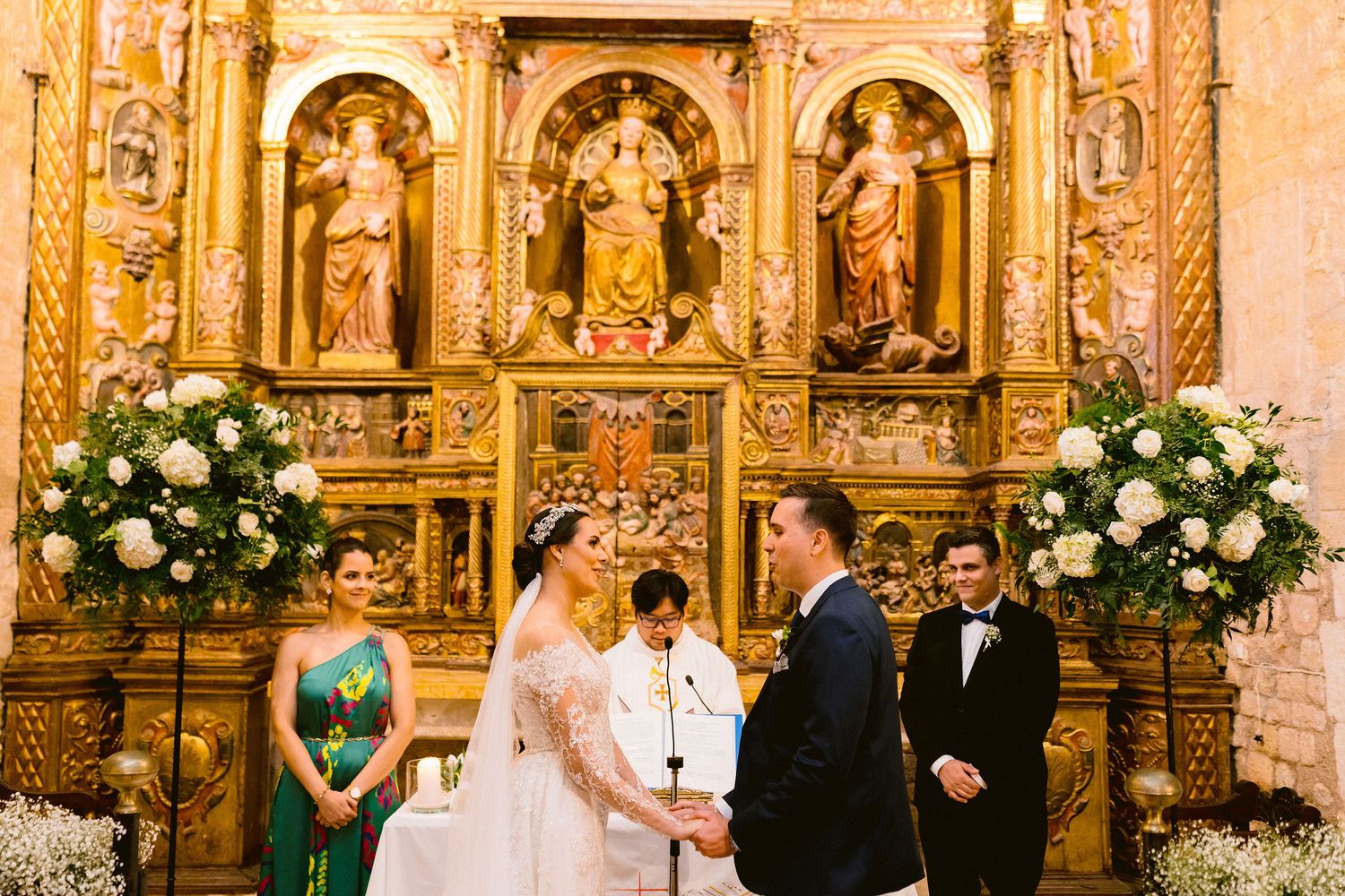 An elegant church wedding ceremony takes place before an ornate golden altar with large white floral arrangements.