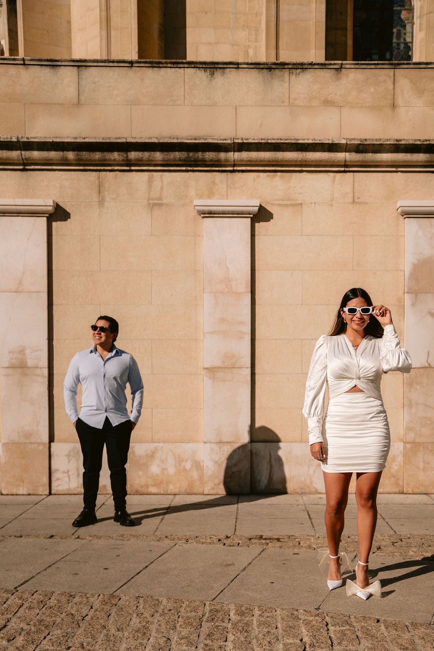 A couple in contrasting outfits pose separately against a beige stone building wall.