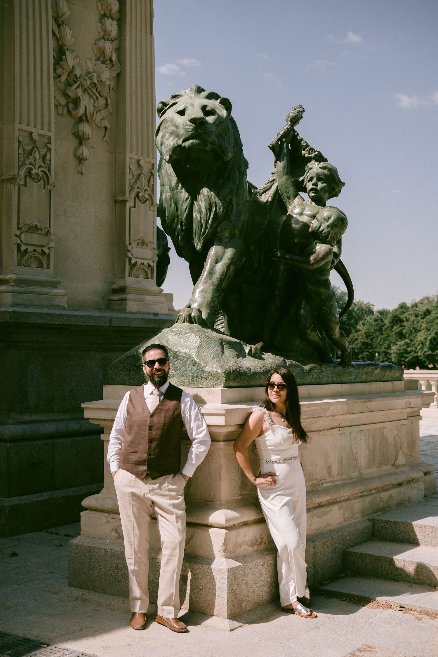 A couple in light colored outfits poses near ornate classical stone sculptures and columns.