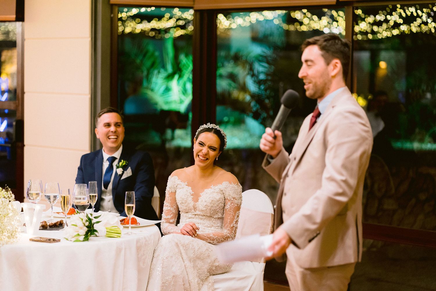 A wedding reception moment at a decorated indoor venue with string lights as a someone gives a speech at the dinner table.