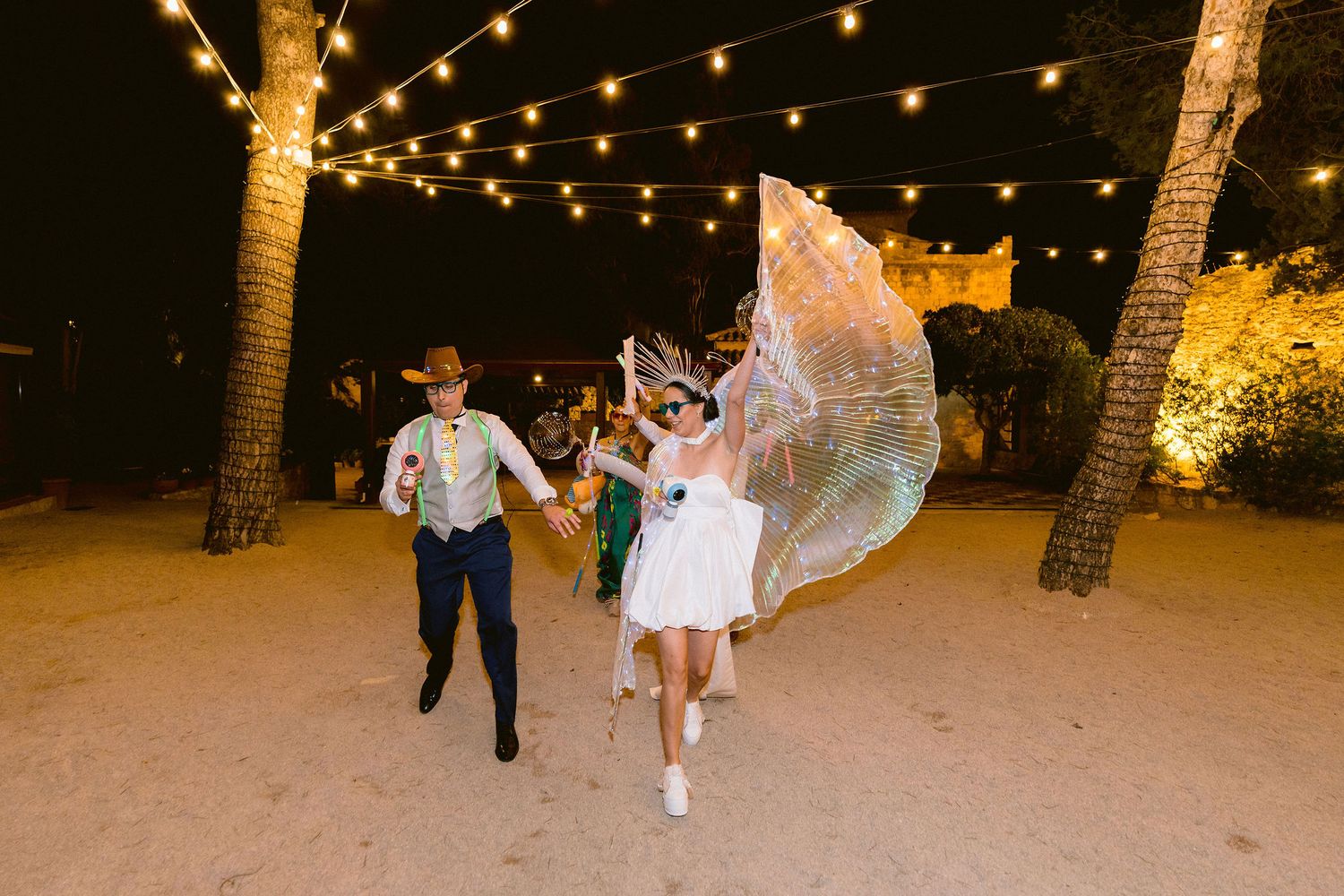 Wedding couple dances on a beach under twinkling string lights and palm trees at night.