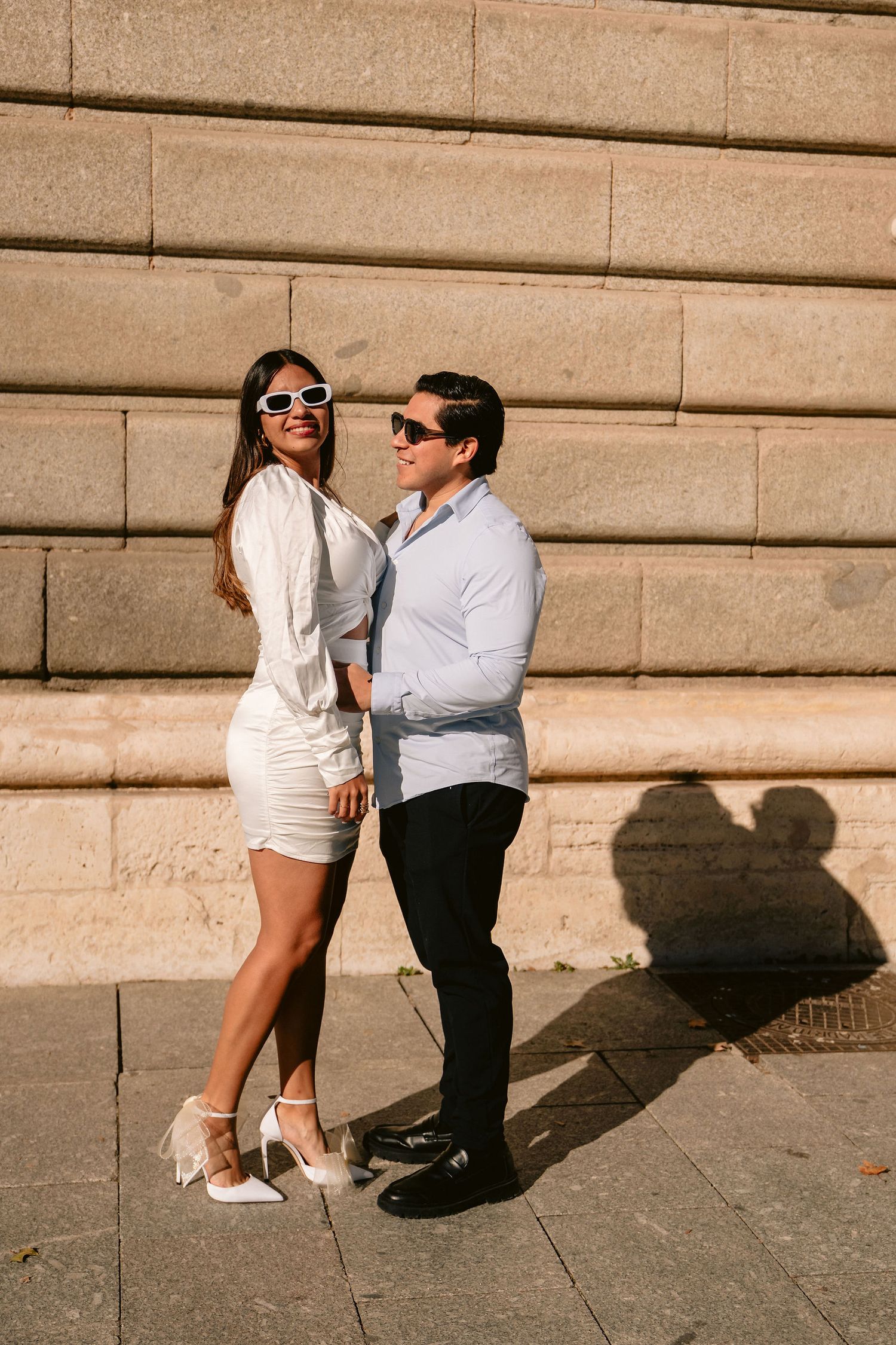 A stylish couple in coordinated white outfits pose against stone steps on a sunny day.
