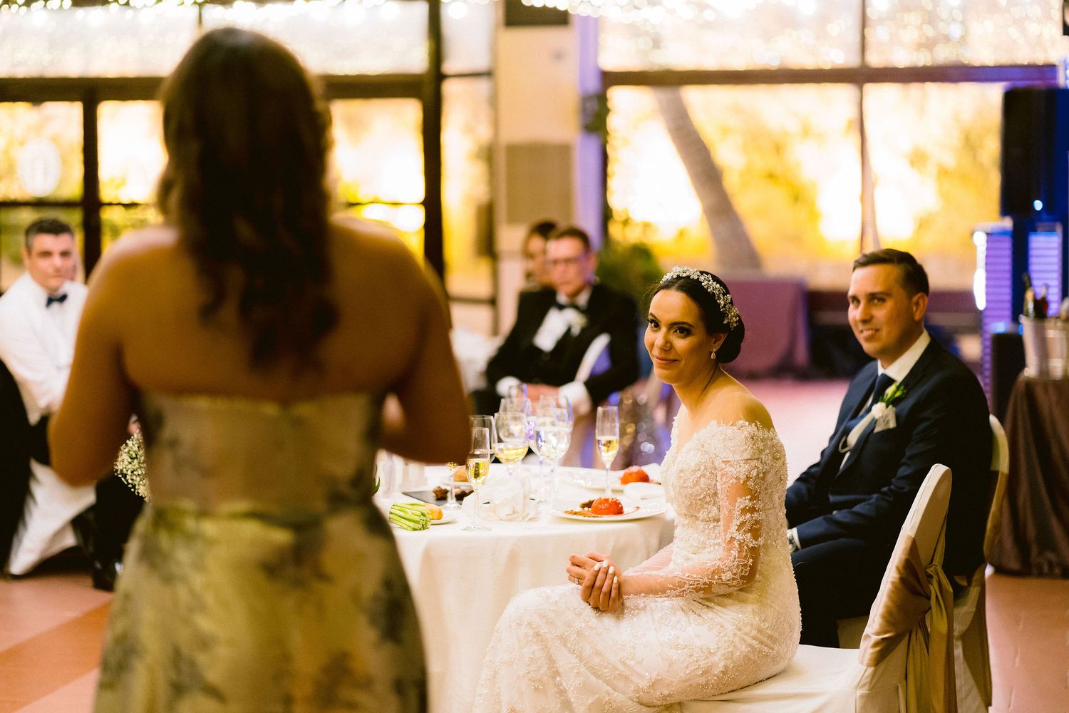 People seated at formal wedding reception tables listen to speech during evening celebration.