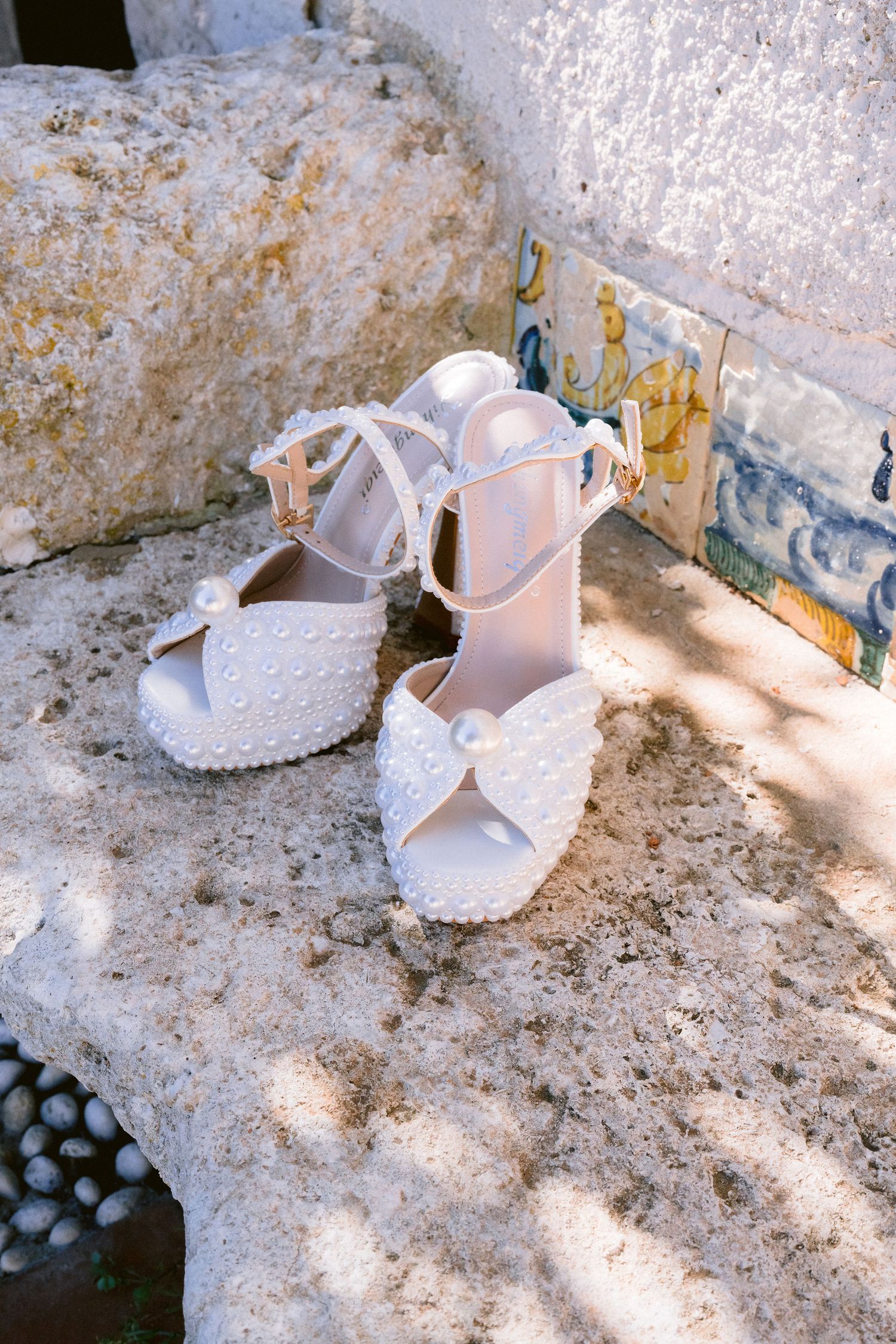 Elegant white lace bridal shoes with ankle straps sit on a rustic stone surface in dappled sunlight.