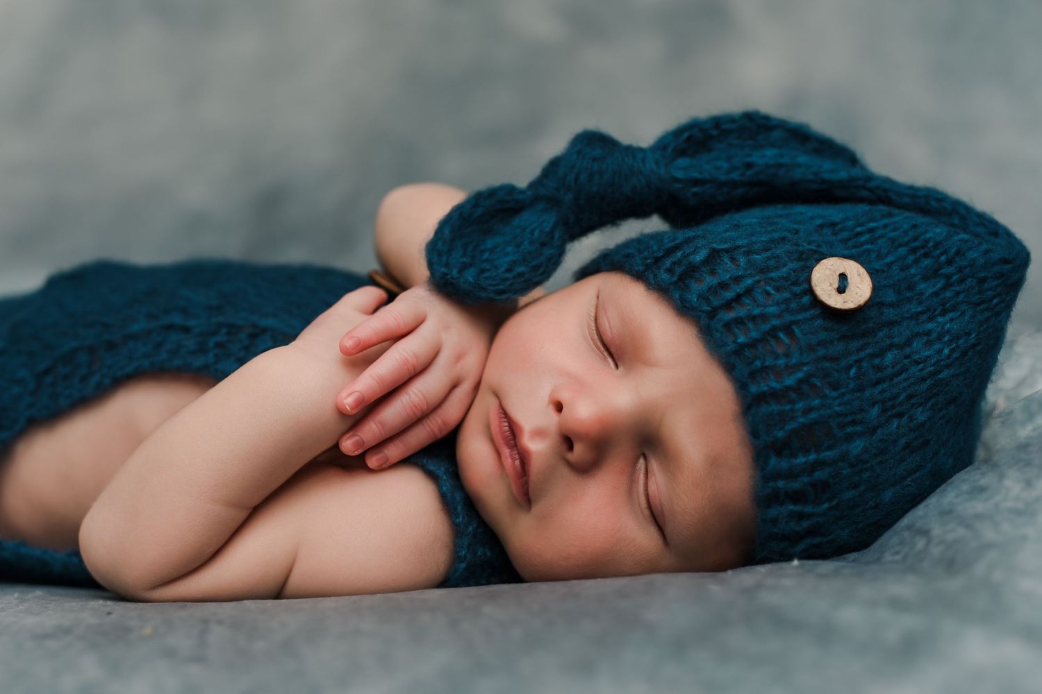 Sleeping newborn baby wrapped in navy blue knit blanket on soft gray backdrop.