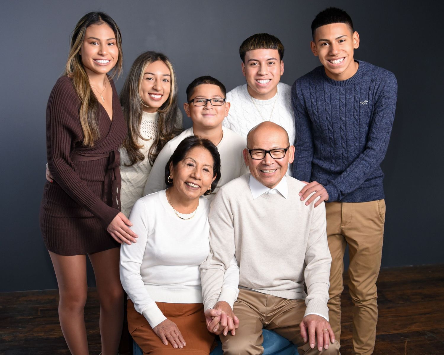 A family group portrait against a dark gray background wearing casual attire in white, brown and blue tones.