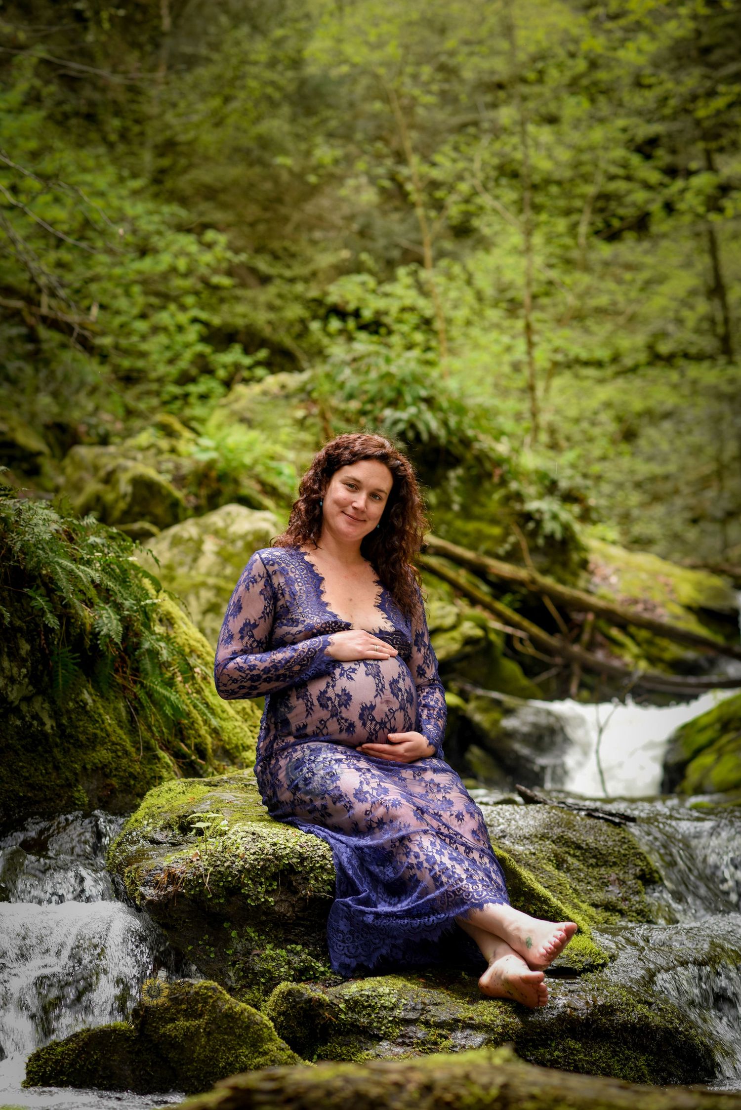 A person in a purple floral dress sits on mossy rocks near a cascading waterfall in a lush forest setting.