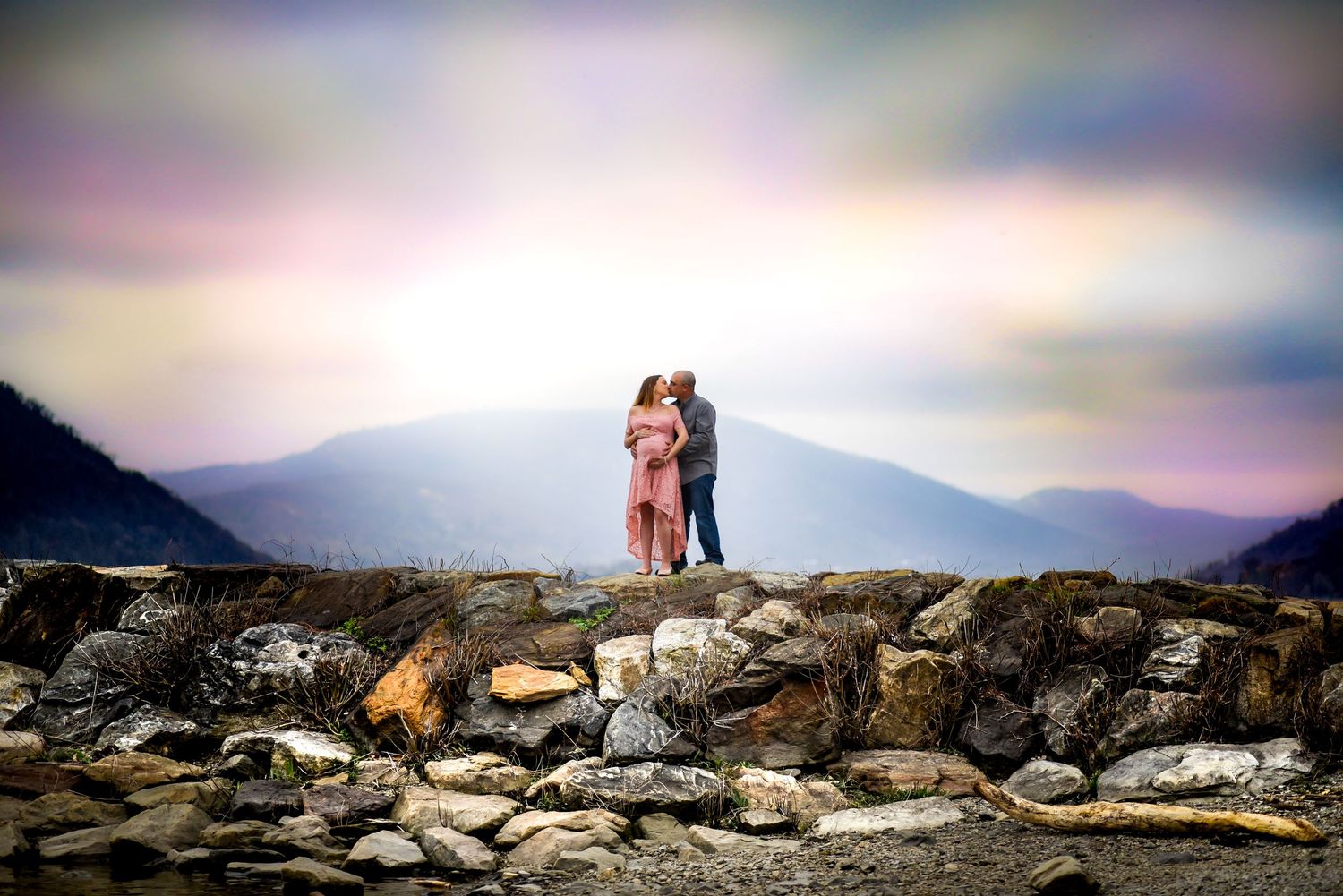 A couple stands on rocky coastal outcrop with misty mountains and colorful sky in the background.