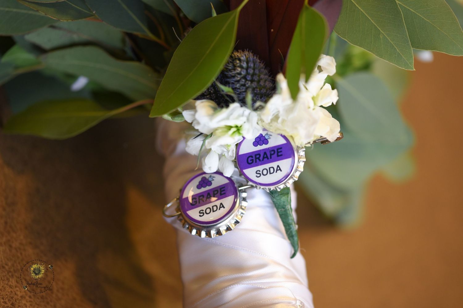 Wedding boutonniere wrapped with purple bottle cap pins against green foliage.