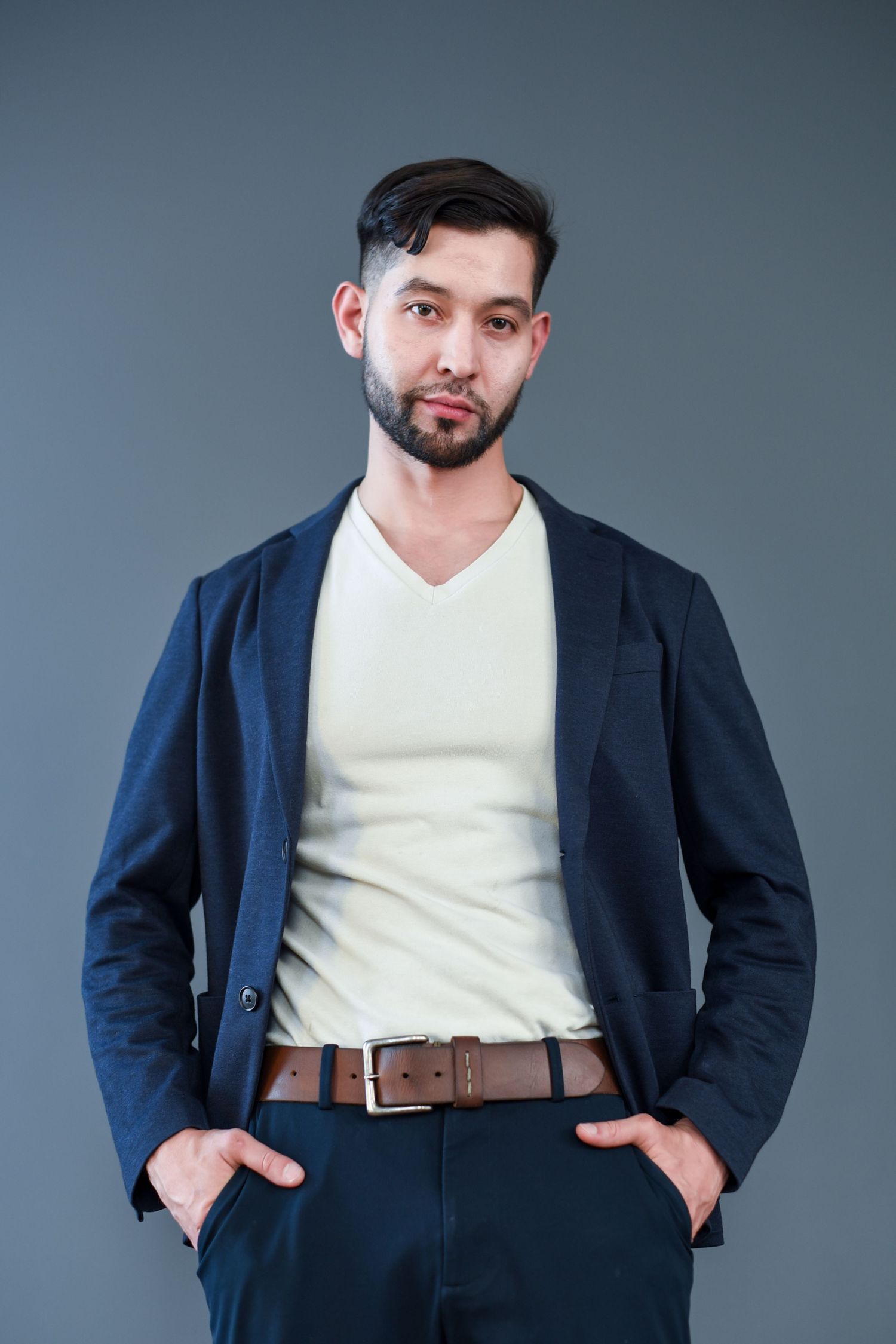 Professional studio portrait showing a navy blazer and white v-neck shirt with hands in pockets against a gray backdrop.