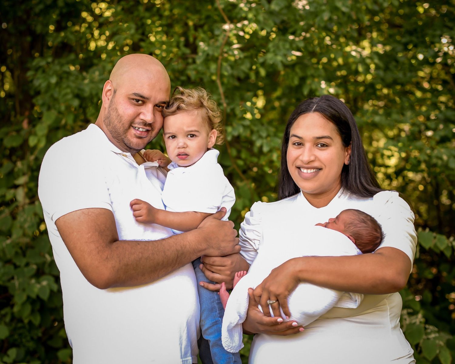 A family in white shirts poses together outdoors against a green leafy background on a sunny day.
