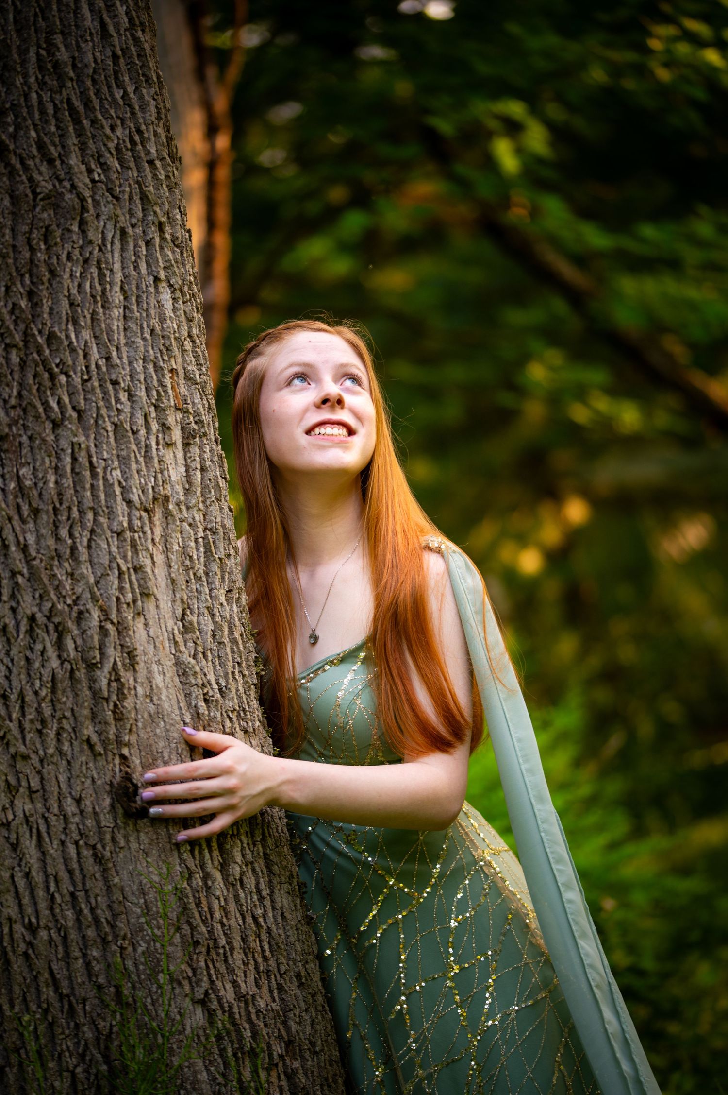 A person in a green medieval style dress poses joyfully against tree trunks in a forest setting.