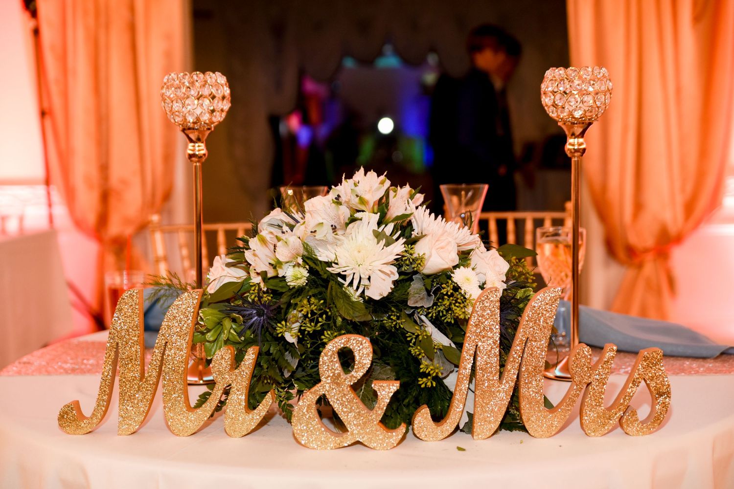 Elegant wedding table decor with 'Mr & Mrs' sign and floral centerpiece in peach lighting.