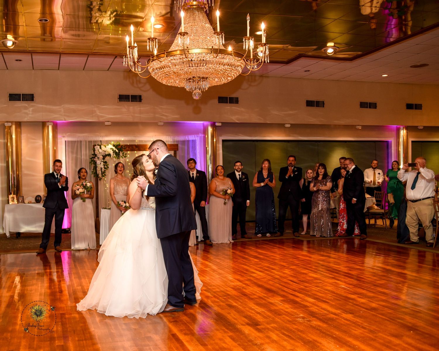 A couple dances in an elegant ballroom with hardwood floors and crystal chandeliers during a wedding reception.