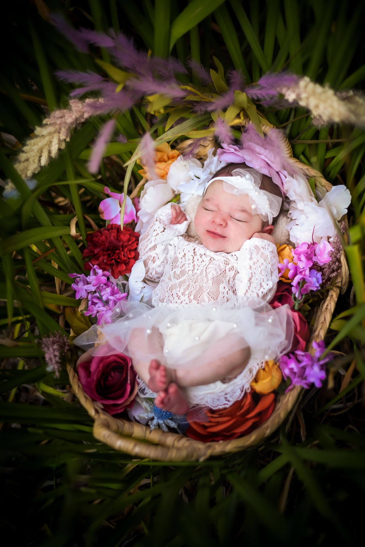 A newborn rests peacefully in a nest-like setting surrounded by delicate pink and purple flowers and natural greenery.