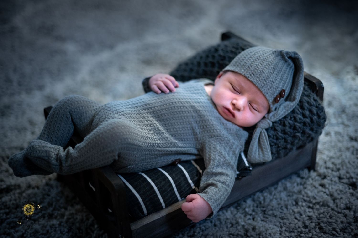A sleeping baby wearing a gray knit outfit and beanie lies peacefully on a dark textured surface.