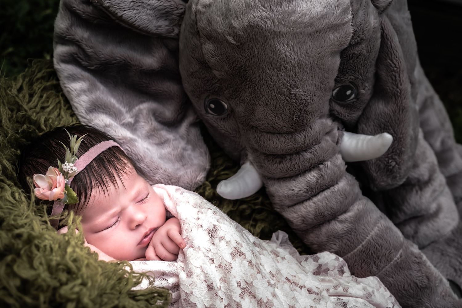 Baby elephant plush toy sits next to sleeping infant in soft pink outfit.