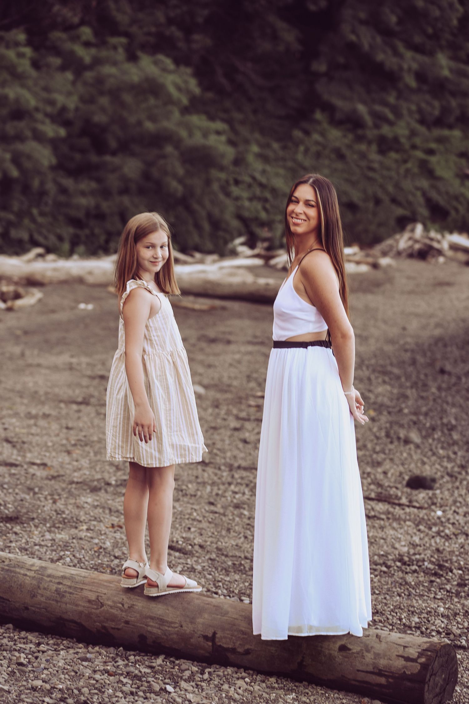 Two people in white dresses stand on a driftwood log at a rocky beach with trees in the background.