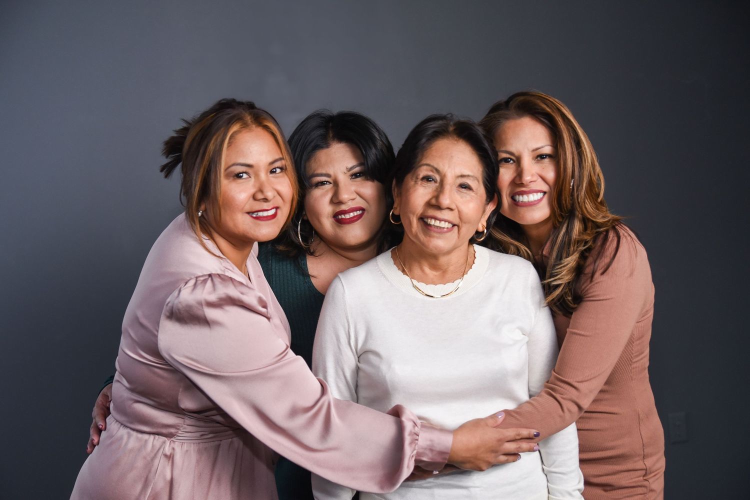 A group of people wearing pink and brown tones embrace and smile together in a studio setting.