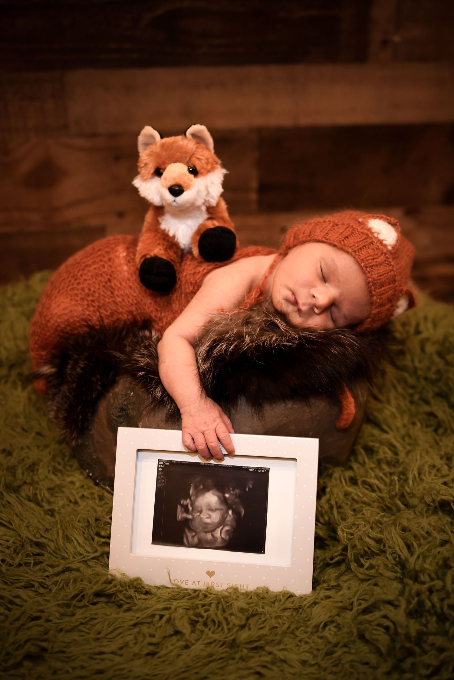 A newborn wearing a fox costume lies on green carpet while holding an ultrasound photo in a creative progression shot.
