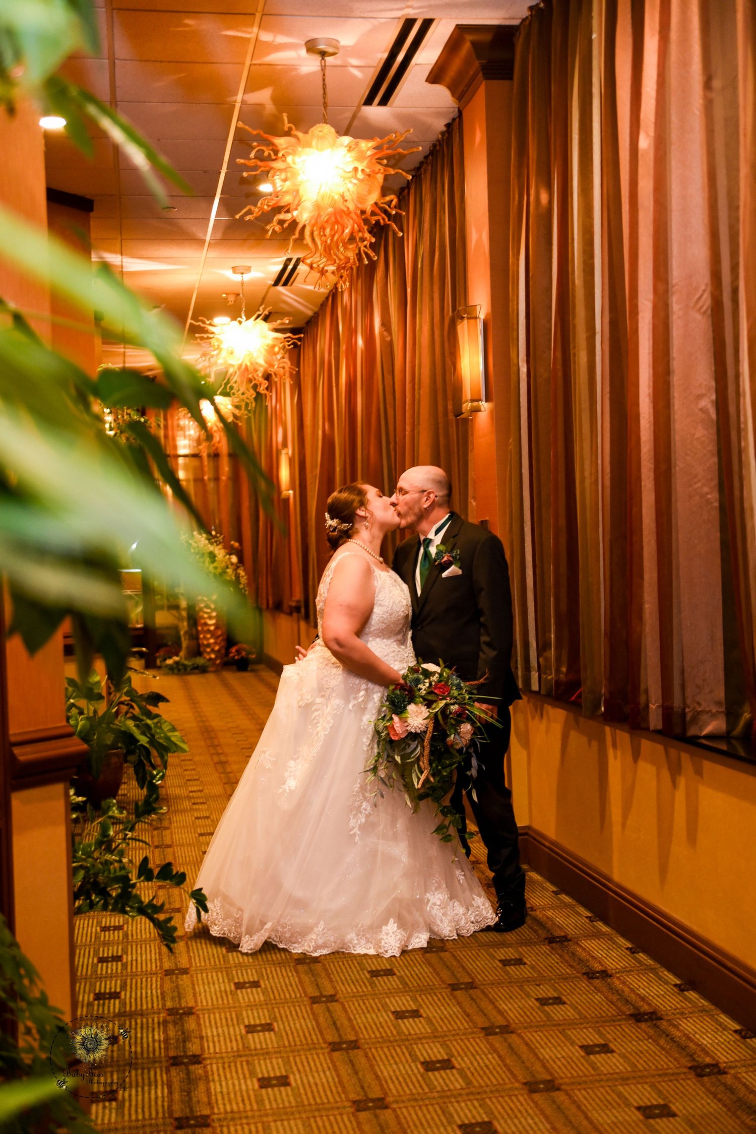 A romantic wedding moment captured in a warmly lit hallway with dramatic drapes and sparkly chandeliers.