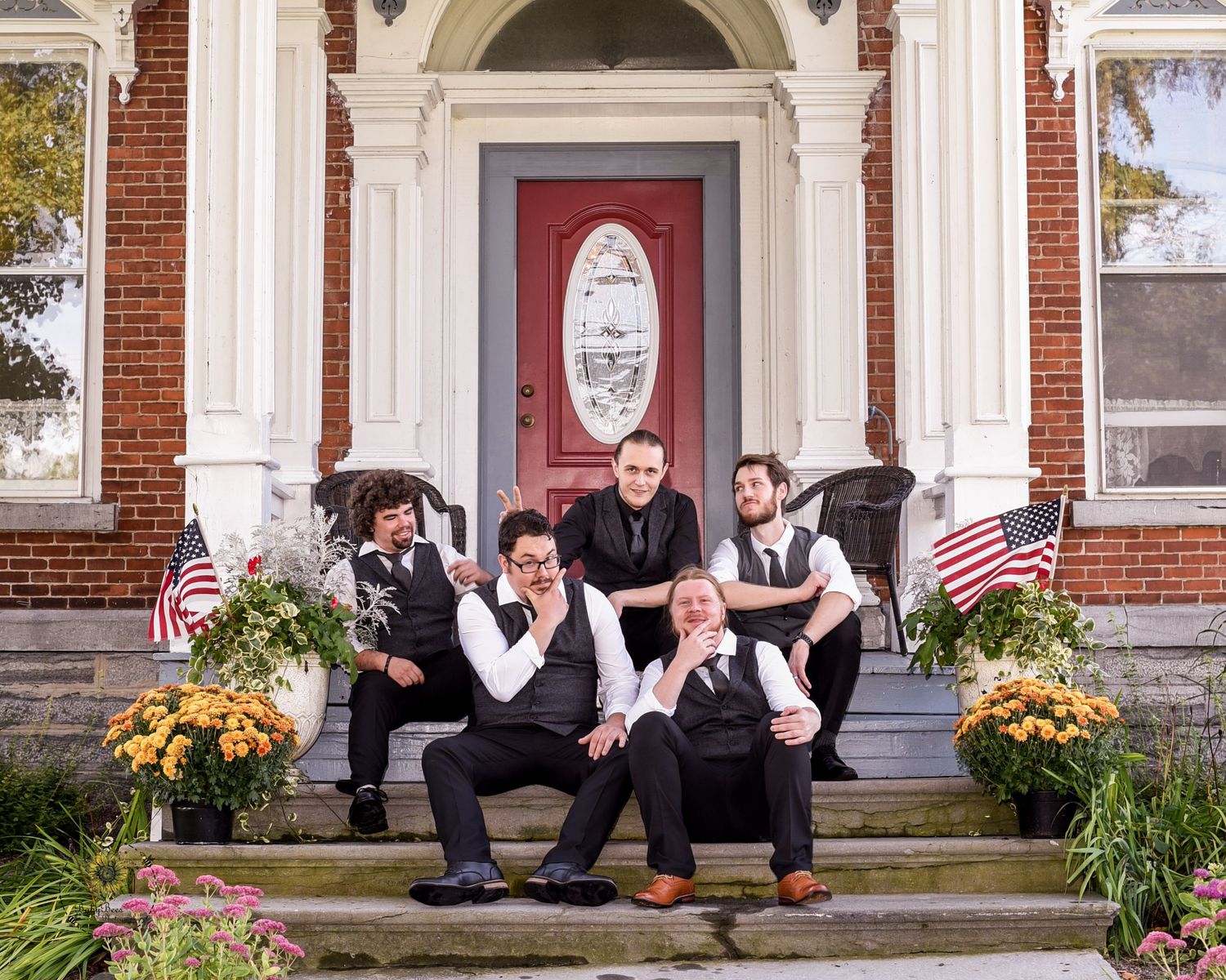 Wedding party poses playfully on brick building steps decorated with autumn flowers and American flags.