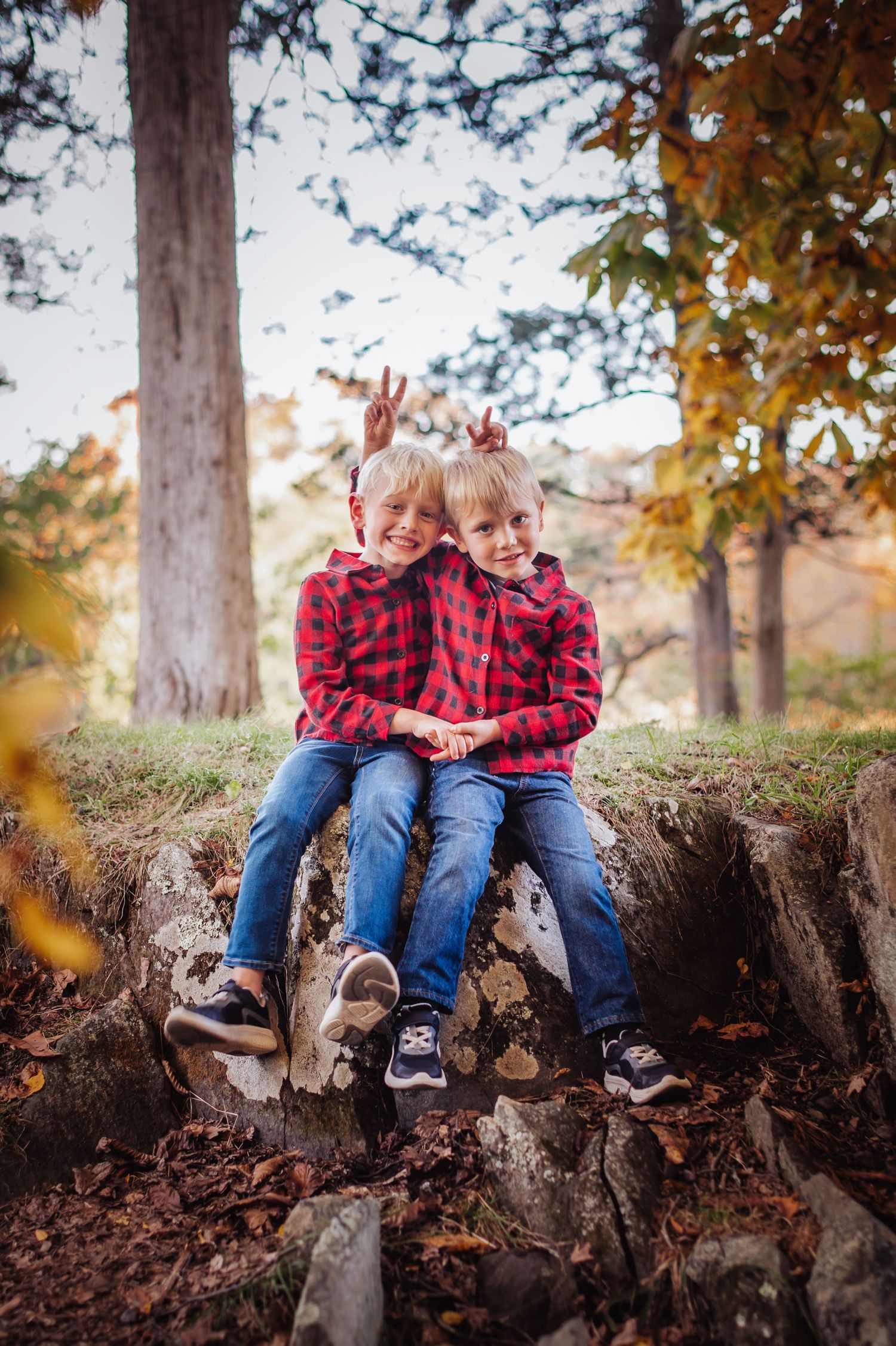 Two children in matching red plaid shirts sit together on tree roots in an autumn forest setting.