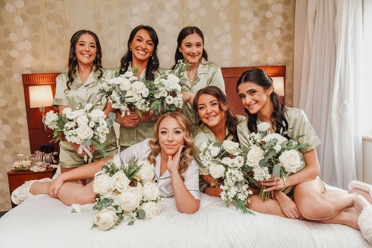Wedding party in white dresses lounging together on a bed with floral bouquets.