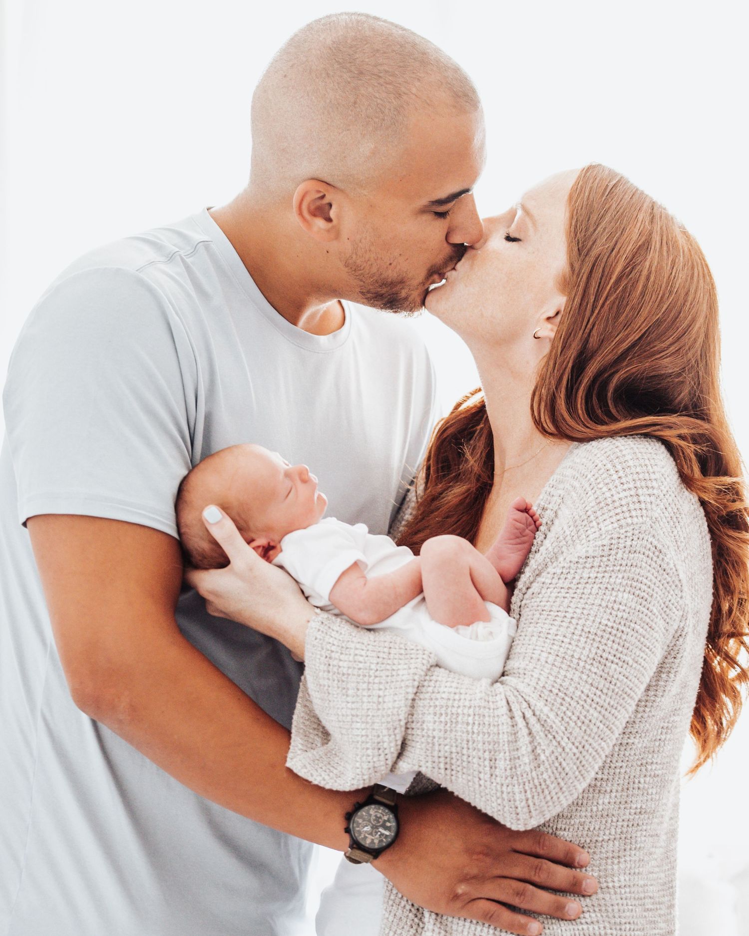 A couple shares a tender kiss while holding their newborn baby dressed in white against a light background.