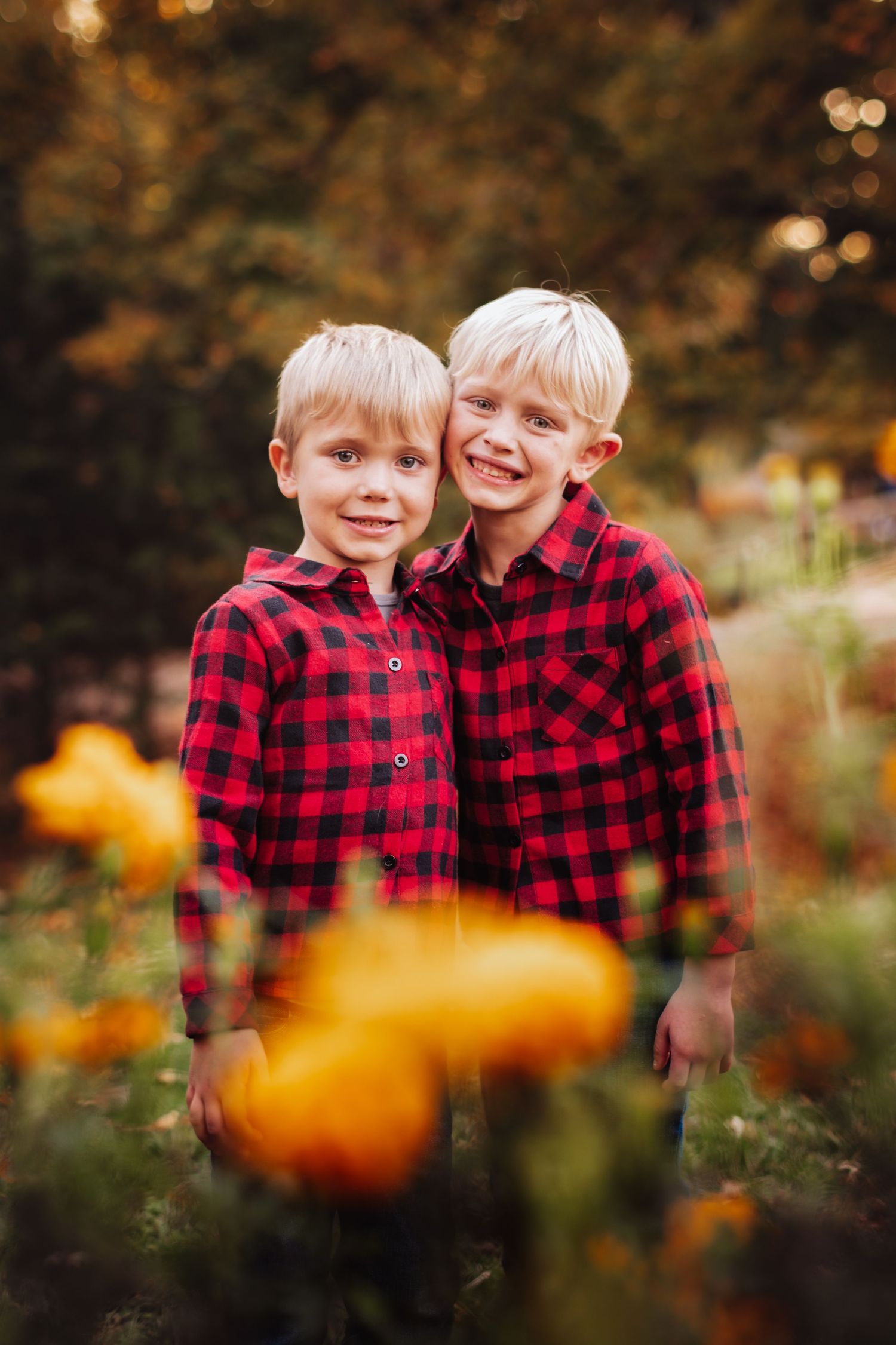 Two children in matching red buffalo plaid shirts share a joyful moment in an autumn garden with pumpkins.