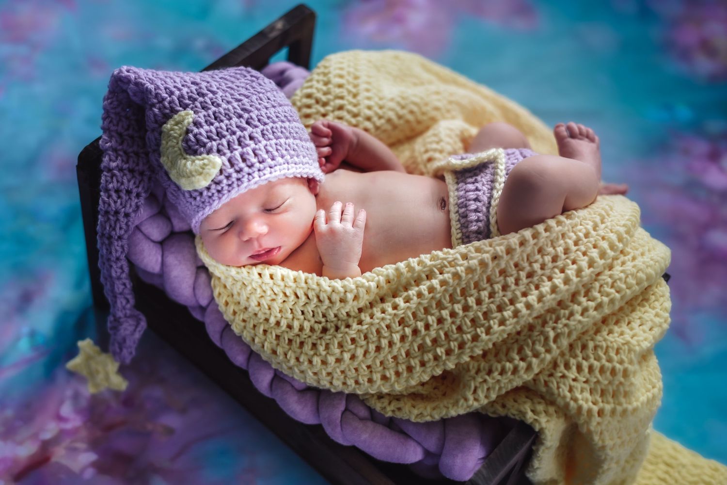 Sleeping newborn wearing yellow and purple crochet outfit rests on colorful tie-dye background.