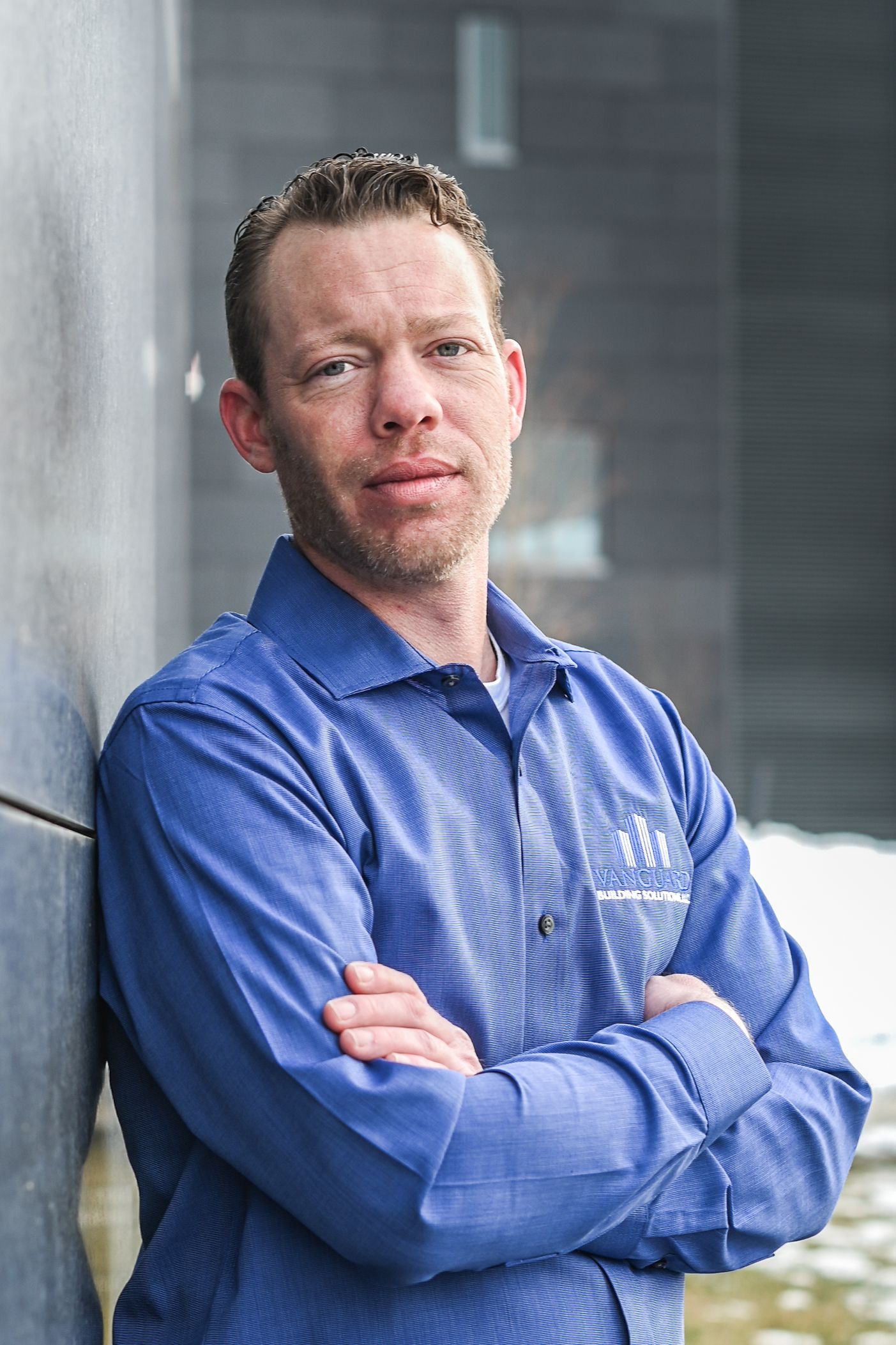 Professional wearing a blue collared uniform shirt stands confidently with arms crossed against a wall.