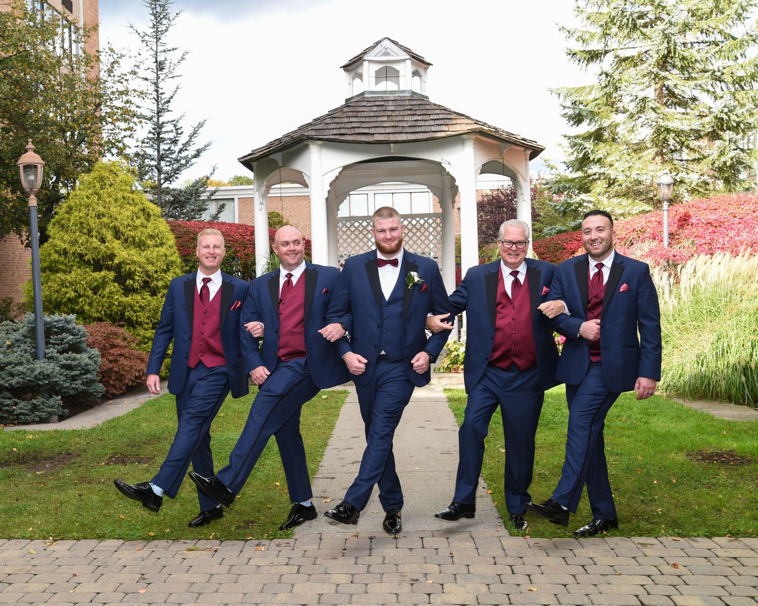 Groomsmen in navy suits and burgundy ties pose playfully on a brick pathway.