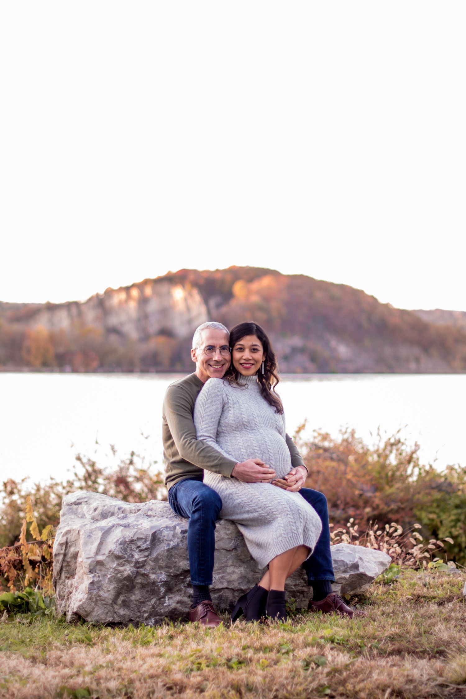 Two people sit together on a large rock in a scenic outdoor setting during autumn.