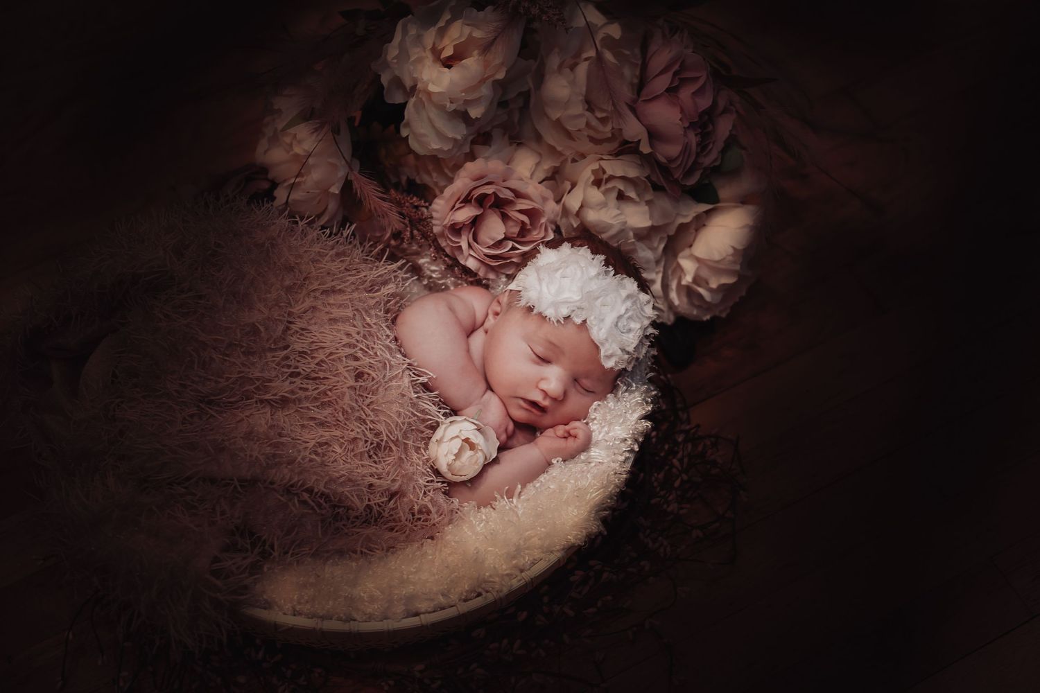 Sleeping newborn in soft pink light surrounded by delicate floral arrangements and textured fabric.
