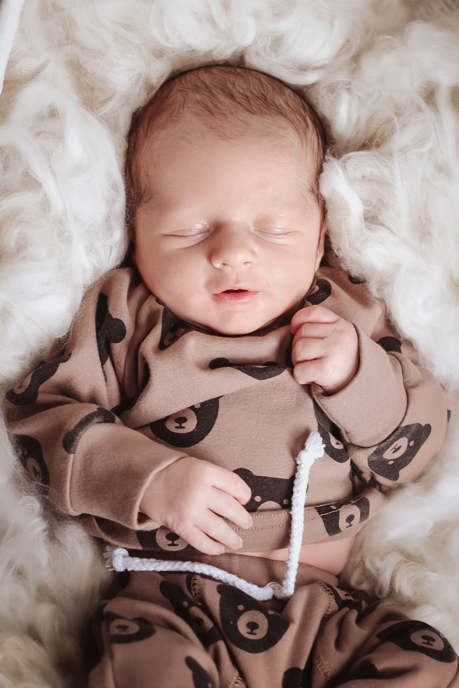 Sleeping newborn baby wrapped in brown patterned blanket lying on soft white fluffy fabric.