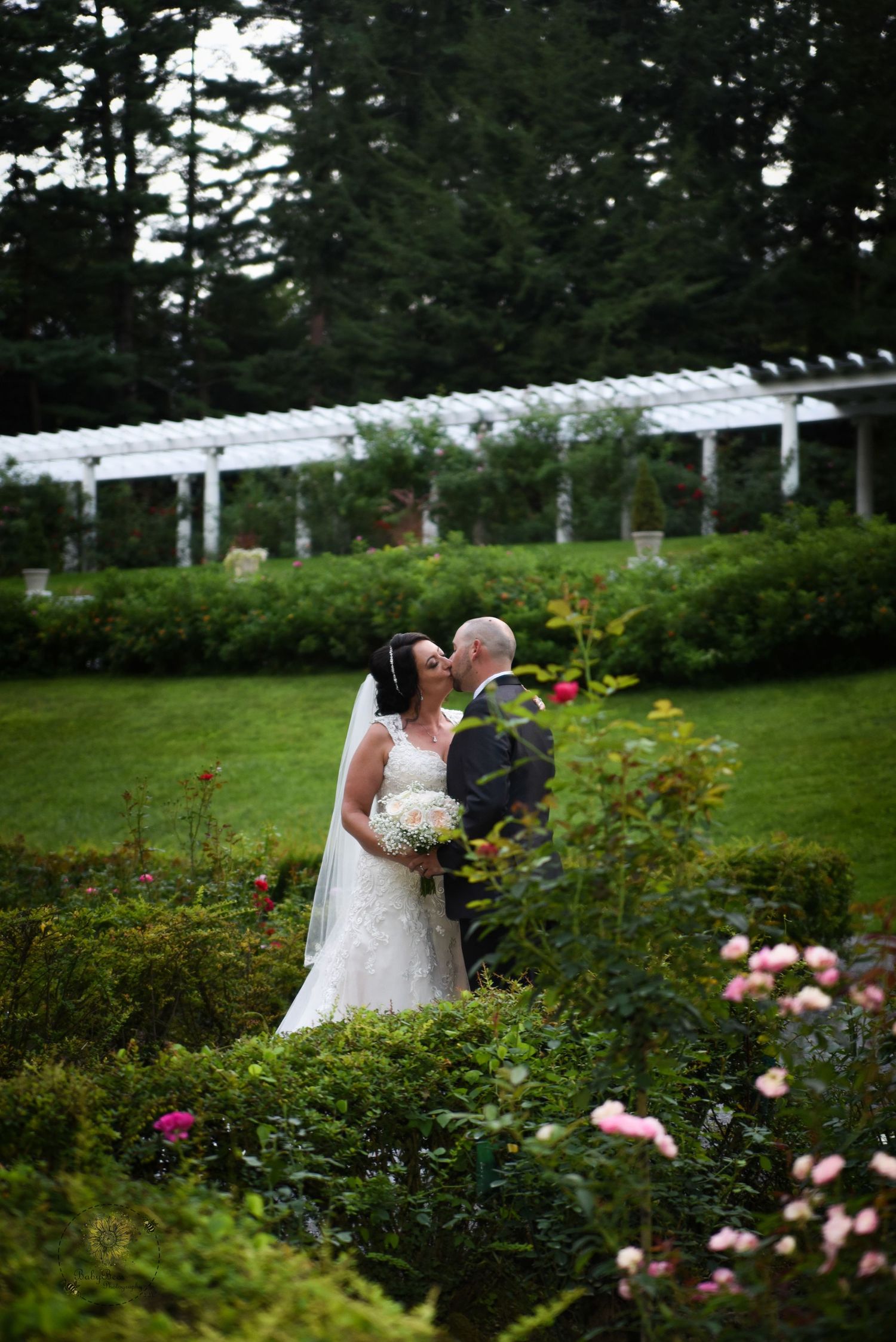 Wedding couple shares intimate moment in lush garden with white pergola and blooming roses.