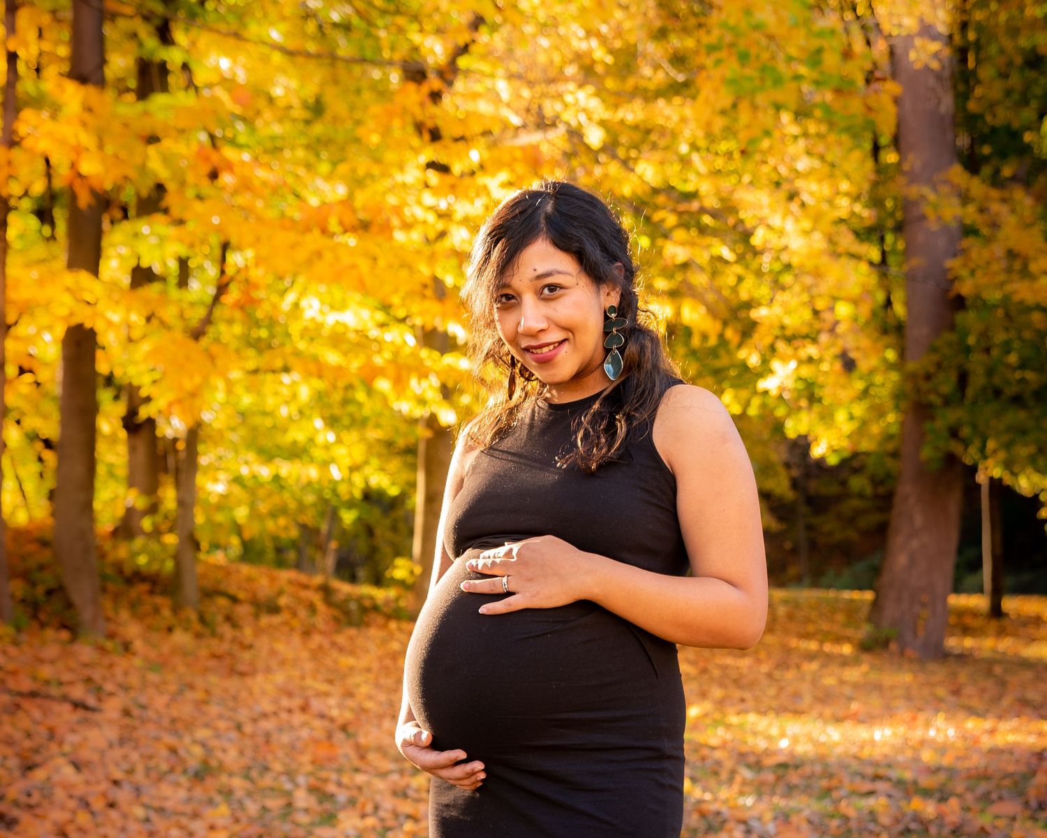 A person in a black dress poses in a vibrant autumn forest setting with golden yellow leaves in the background.