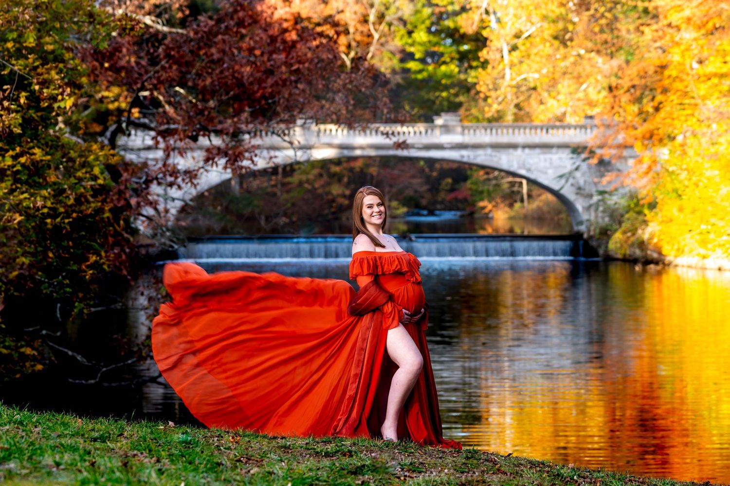 Flowing red dress creates dramatic movement against autumn foliage and reflective water in scenic outdoor photoshoot.