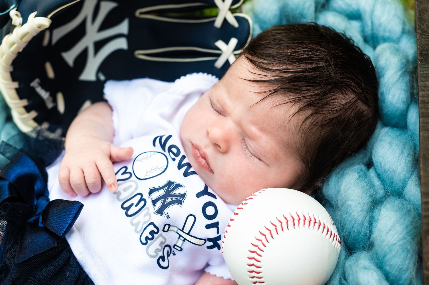 A sleeping baby rests peacefully on a soft blue blanket while holding a baseball.