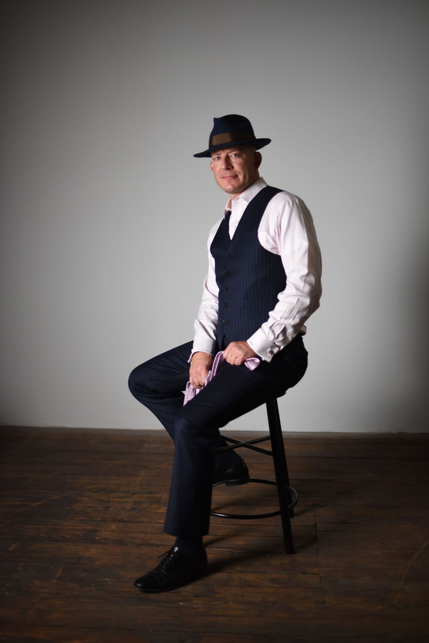 Dramatic portrait series of a person in formal attire with vest and hat seated on dark wooden stool against grey backdrop.