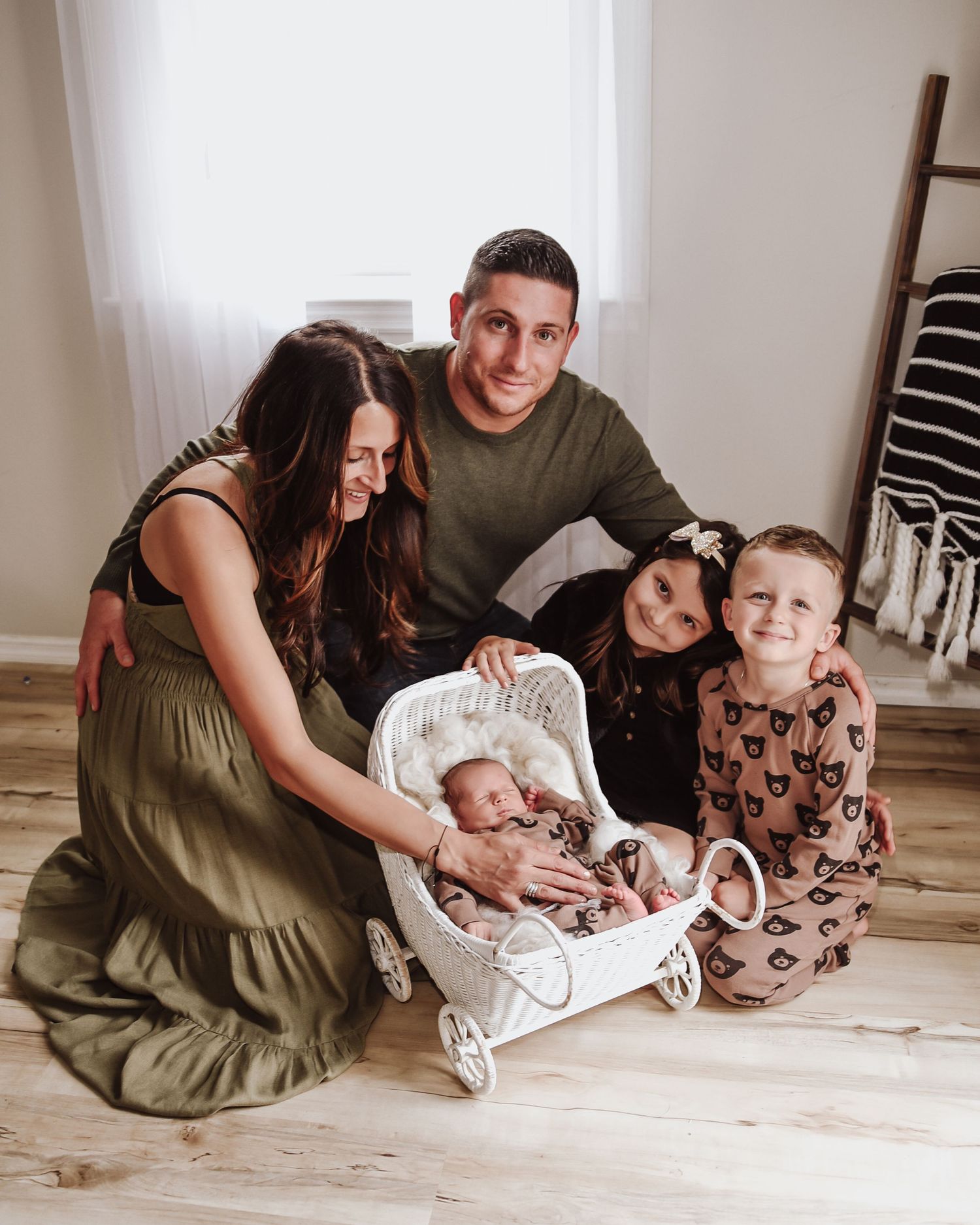 Family poses together on wooden floor with baby in white bassinet for lifestyle photography session.