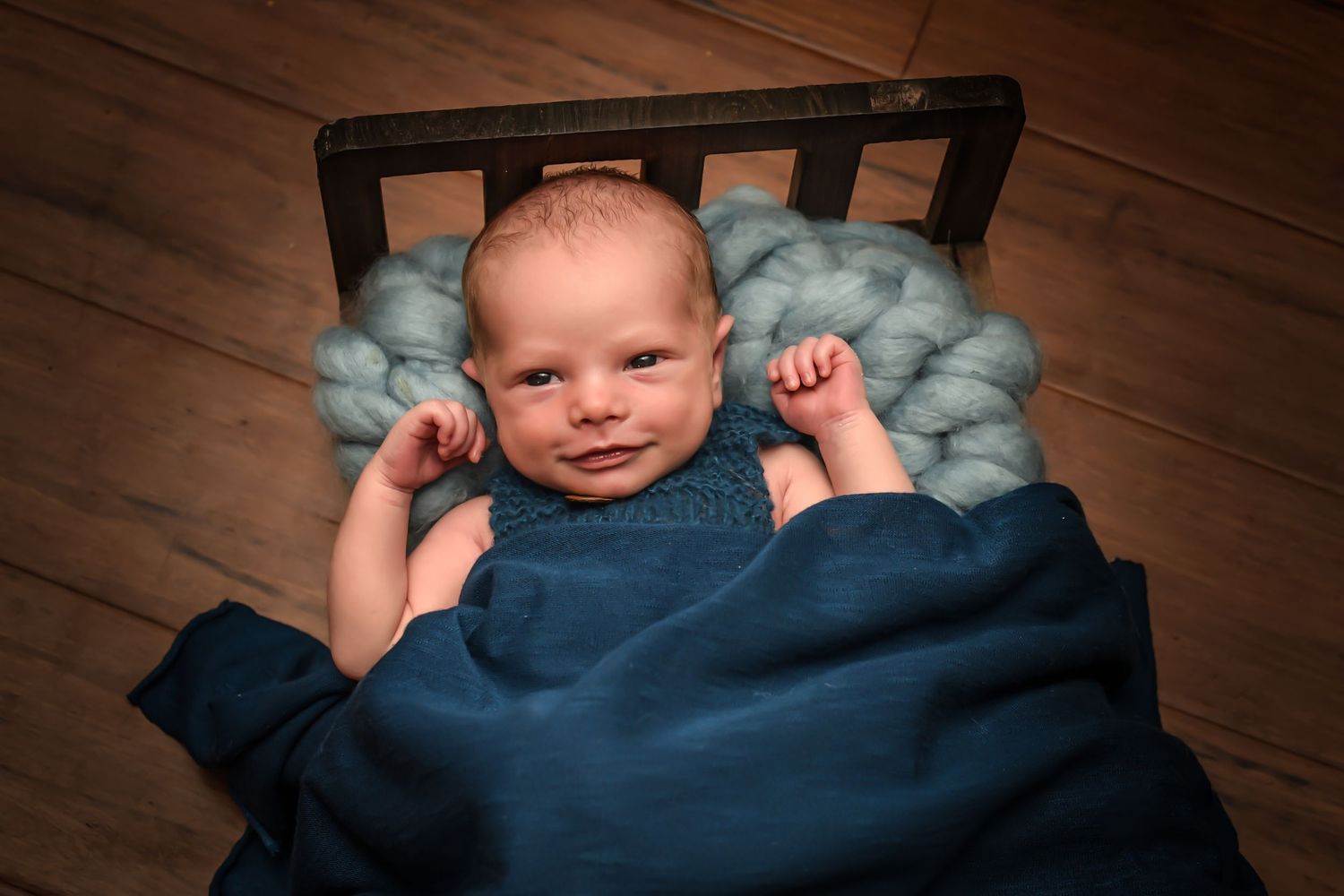 A baby lies in a wooden crate with a navy blue blanket and light blue cushioning on a hardwood floor.