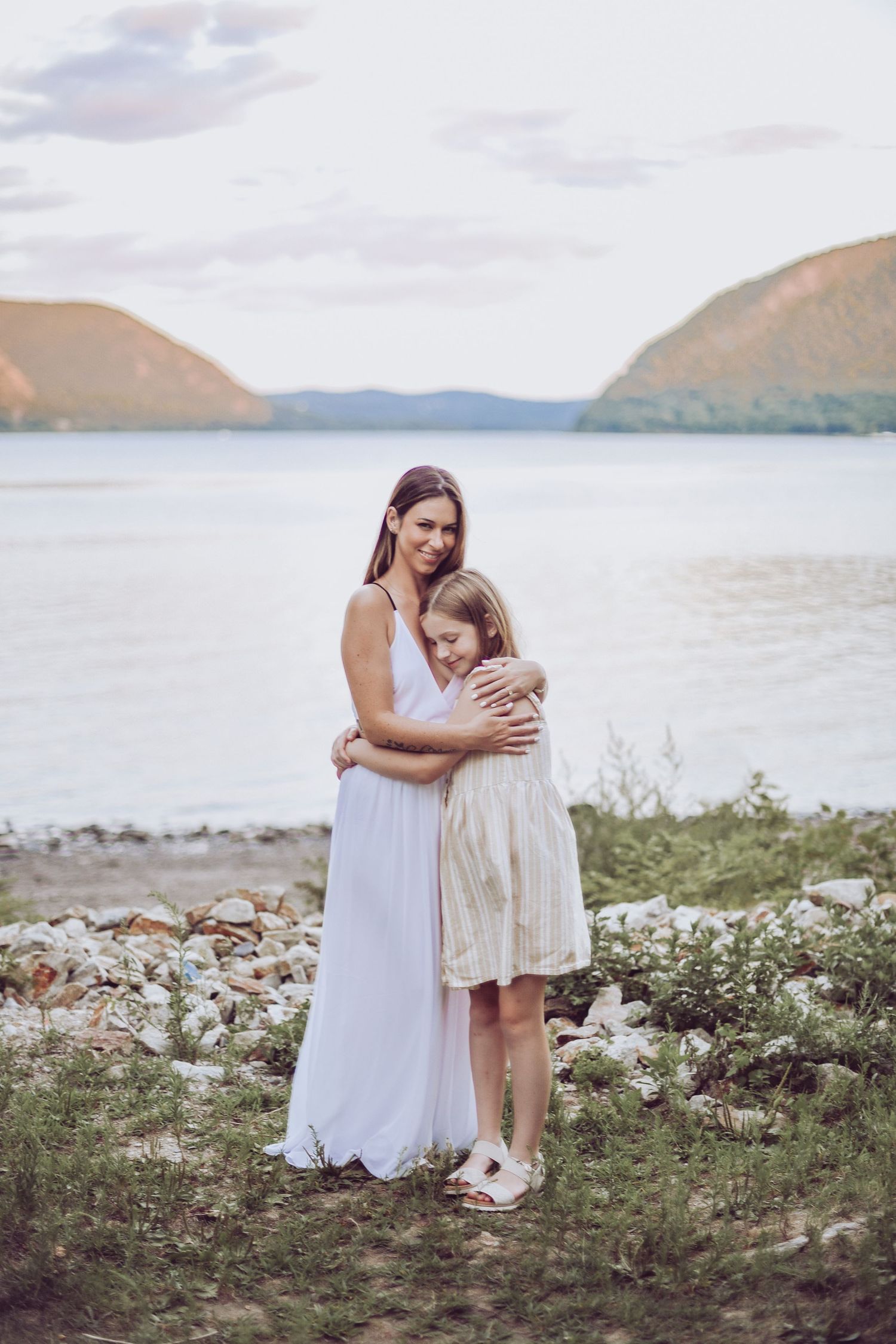 Two people in white dresses embrace while standing on rocky shoreline with mountains and lake in background.